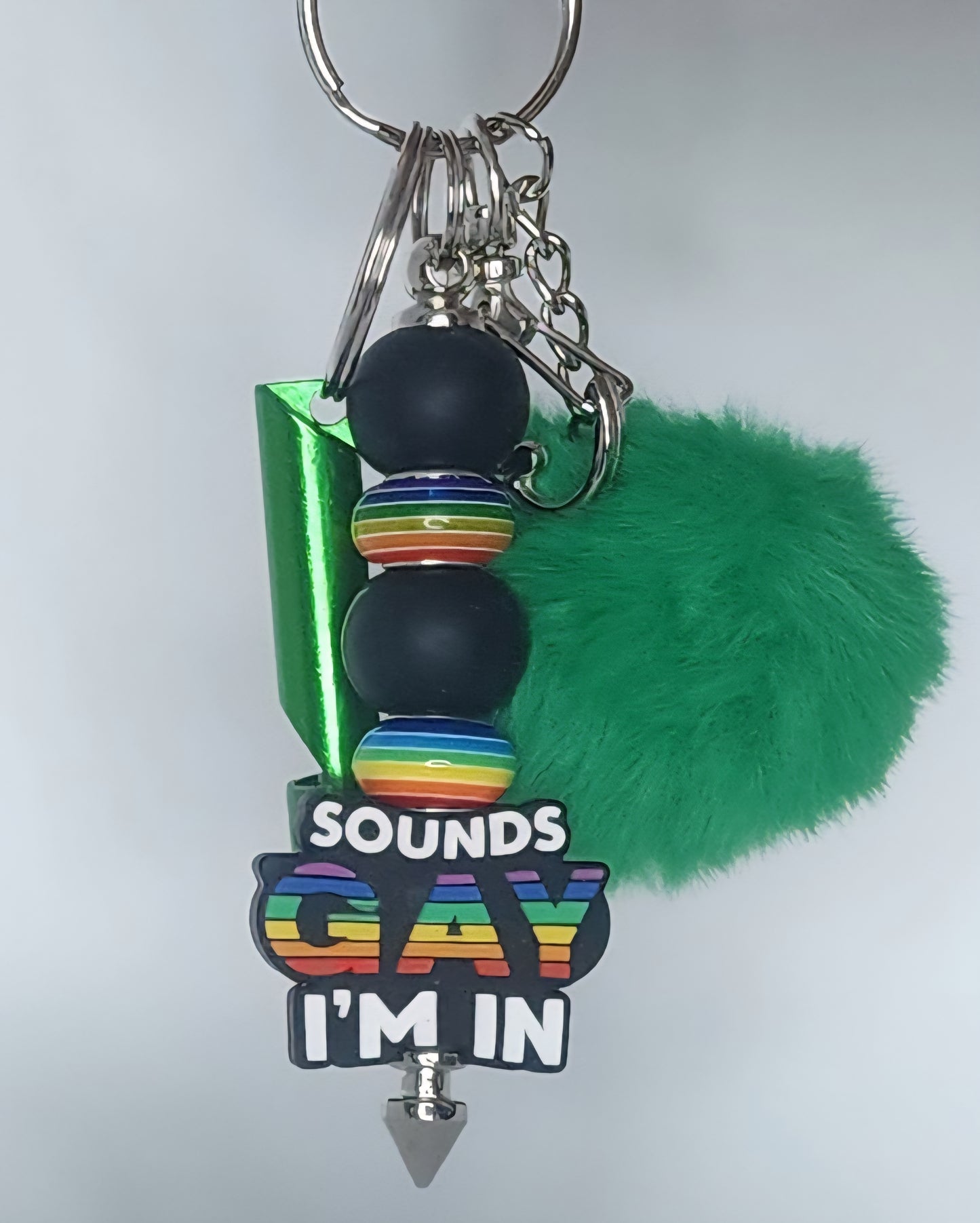 Keychain with rainbow text 'SOUNDS GAY I'M IN' and green fluffy ball on a white background