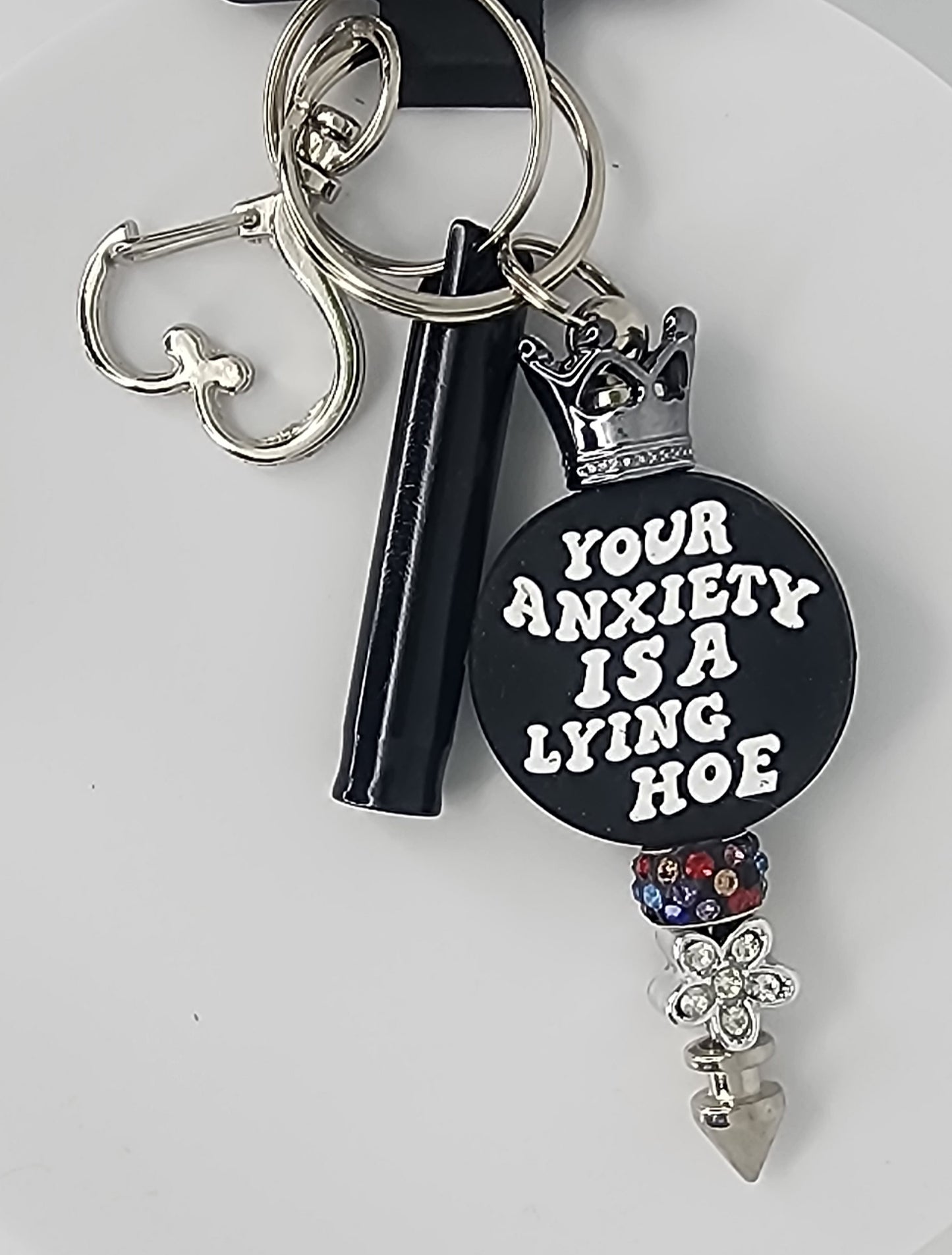 Keychain with a crown and floral bead, and a black bead that says Your Anxiety is a Lying Hoe, with a black whistle and heart shaped clip on a white background.
