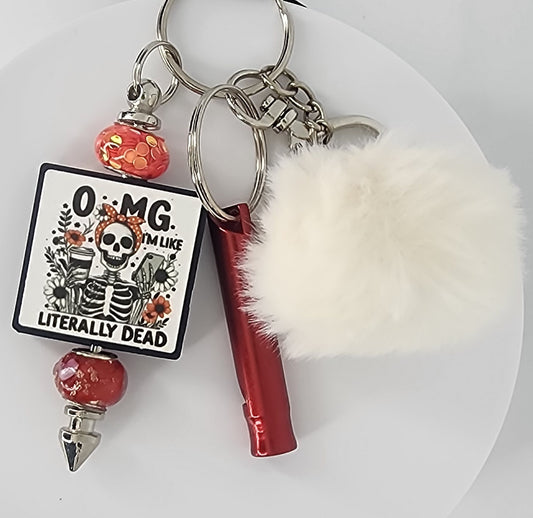 Keychain with skeleton design that says OMG. I'm Like Literally Dead, red safety whistle, and pom-pom on a white background