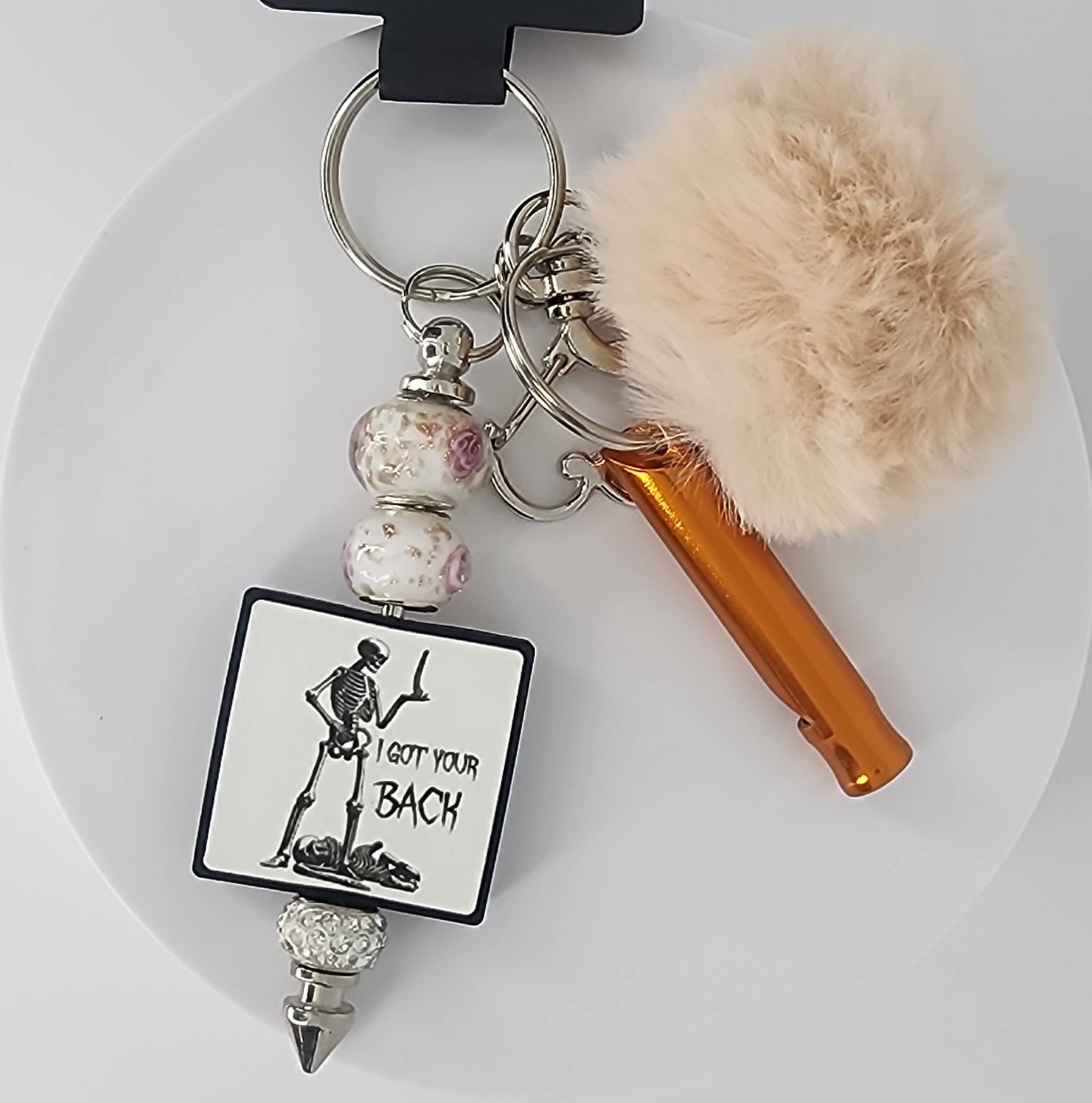 Keychain with skeleton design that says "I got your back" with a whistle and pom-pom on a white background