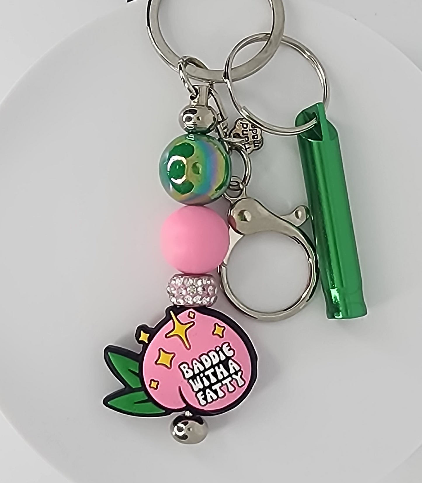 Peach shaped silicone bead that says "Baddie with a Fatty" on a keychain with matching pink and green accent beads, a green whistle and a clip.