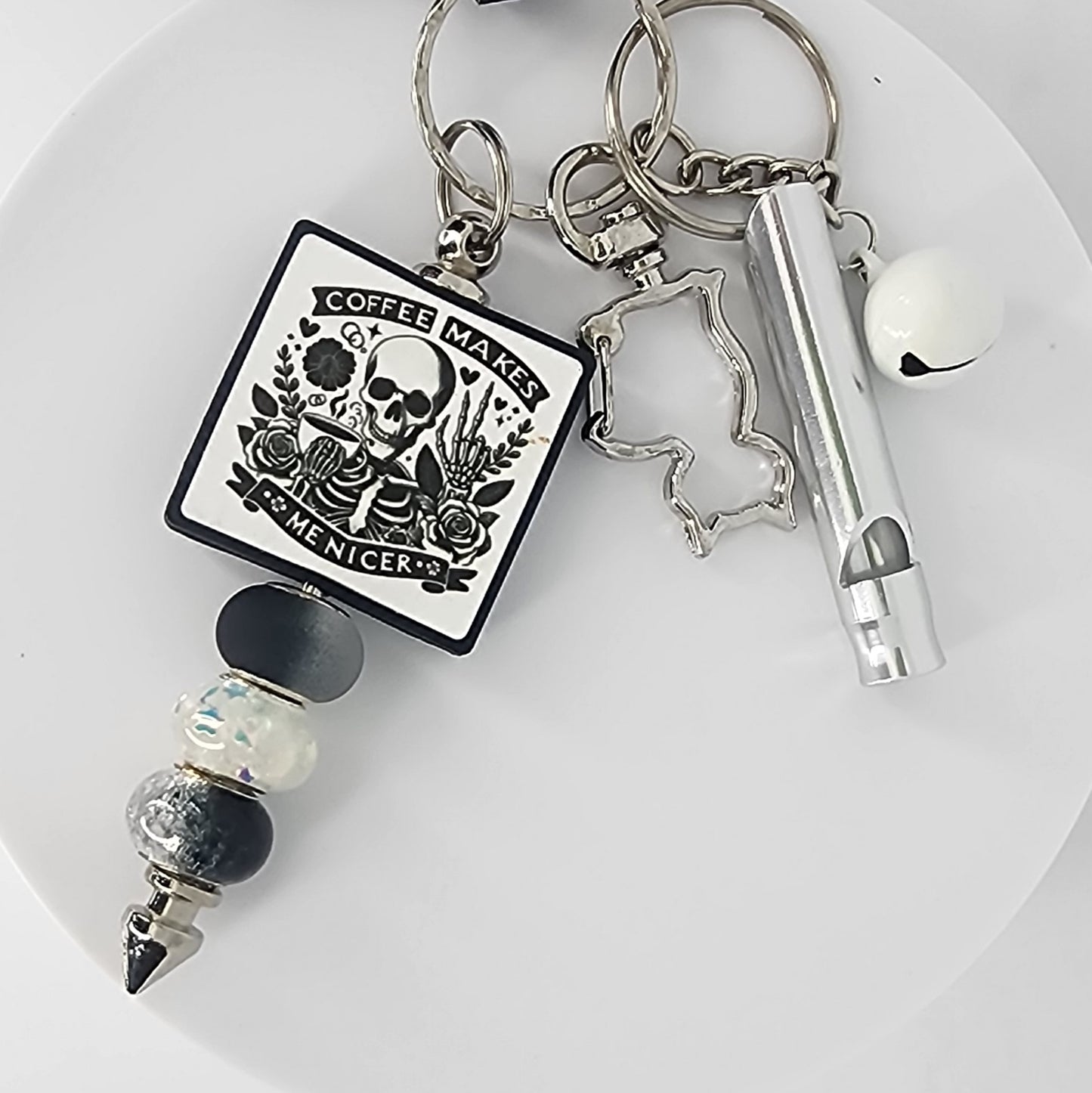 Keychain with a skeleton design that says "Coffee Makes Me Nicer," a cat-shaped clip, a white jingle bell, and a silver safety whistle.